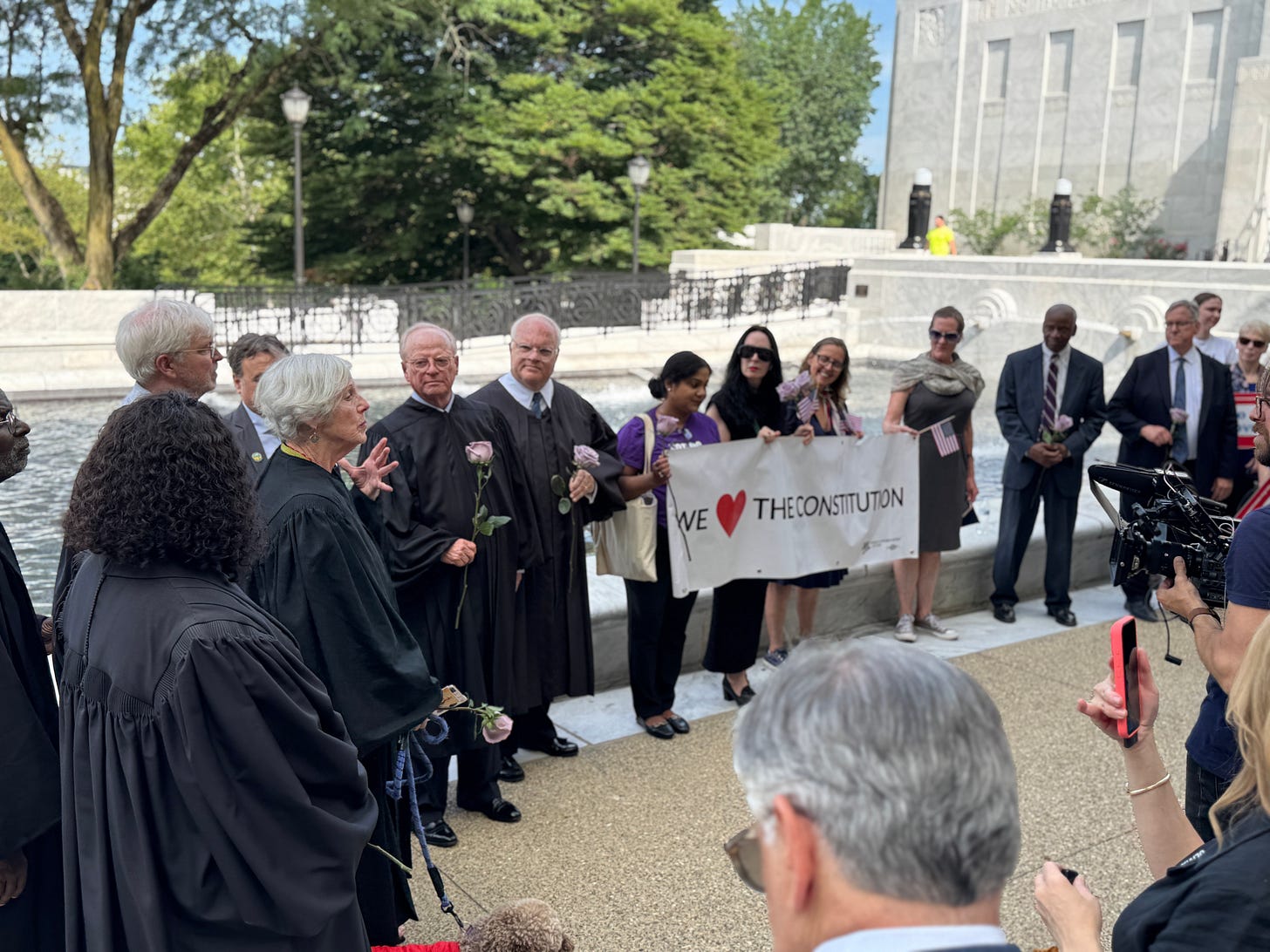 an outdoor photo in front of a courthouse. on the left, a group of male and female judges, one speaking. on the right, a group of people holding a big banner reading "we <3 the constitution" an outdoor photo in front of a courthouse. on the left, a group of male and female judges, one speaking. on the right, a group of people holding a big banner reading "we <3 the constitution"