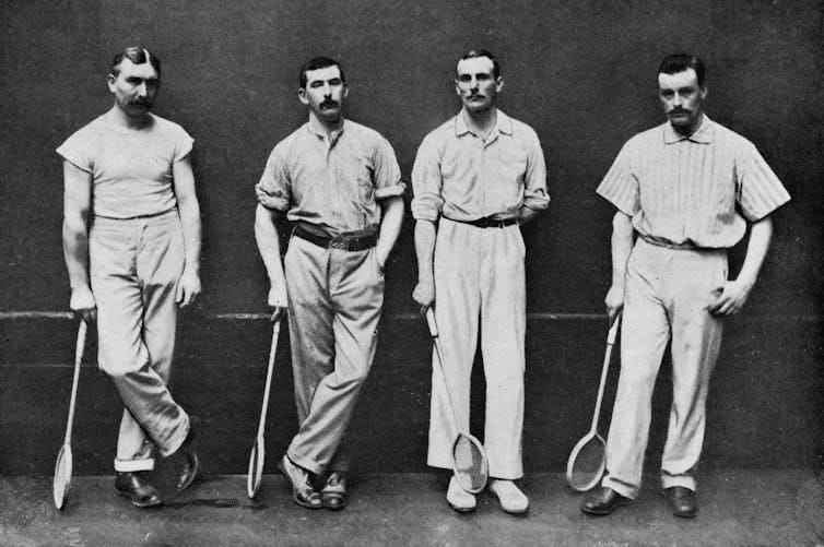 Black and white photo of four men in shirts, slacks and shoes holding squash rackets