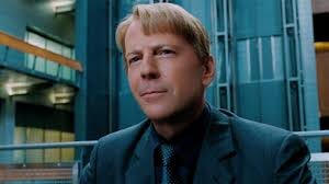 bruce willis, do busto pra cima, de terno cinza azulado, no que parece um escritório no mesmo tom que seu terno. Bruce Willis é um homem branco e loiro, com nariz forte e uma expressão de meio sorriso. Sua pele é notavelmente lisa, de forma quase artificial. Seu cabelo loiro é cortado com uma franja meio galã