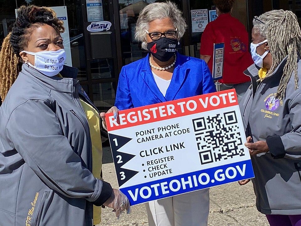 three African American women in masks outside a business, one holding a sign that says register to vote with directions to use a QR code three African American women in masks outside a business, one holding a sign that says register to vote with directions to use a QR code