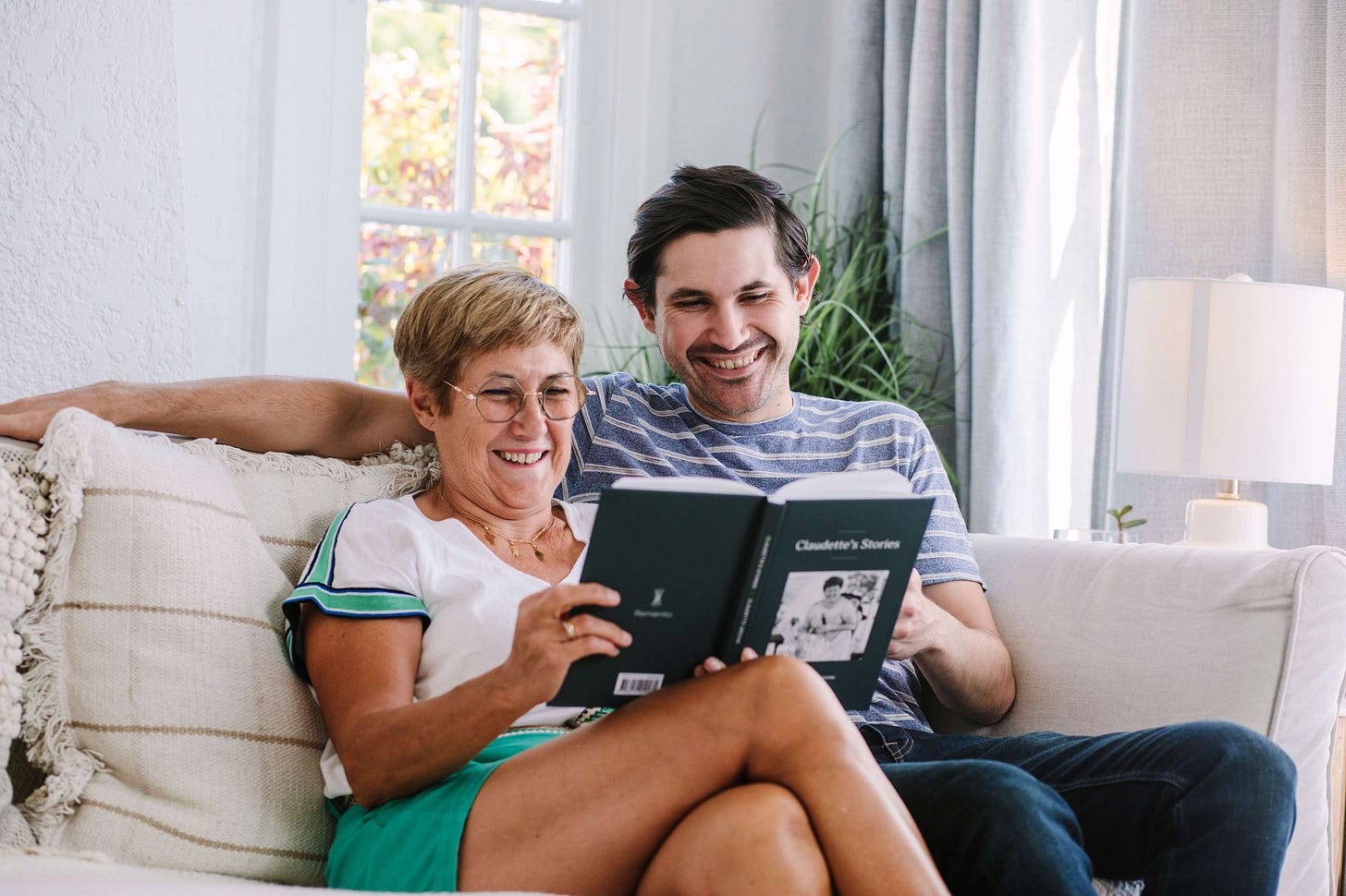 An adult male and his mother sitting on a couch smiling and reading their Remento keepsake book together