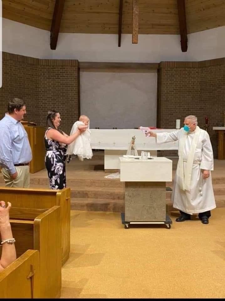 A priest in a white robe stands at an altar, extending a hand toward a baby dressed in a white gown held by a woman in a floral dress. A man in a blue shirt and khaki pants stands beside them. The setting is a church with wooden pews, a brick wall, and a high wooden ceiling. A priest in a white robe stands at an altar, extending a hand toward a baby dressed in a white gown held by a woman in a floral dress. A man in a blue shirt and khaki pants stands beside them. The setting is a church with wooden pews, a brick wall, and a high wooden ceiling.