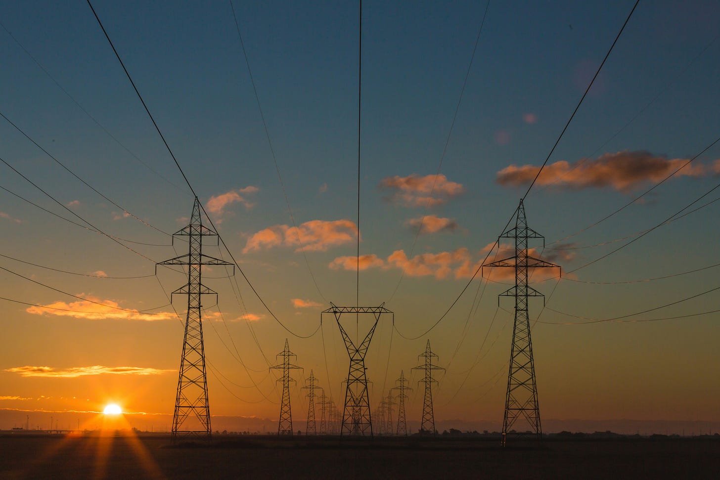 A row of tall metal transmission towers and overhead power lines stretching into the distance against a bright sunset sky. A row of tall metal transmission towers and overhead power lines stretching into the distance against a bright sunset sky.
