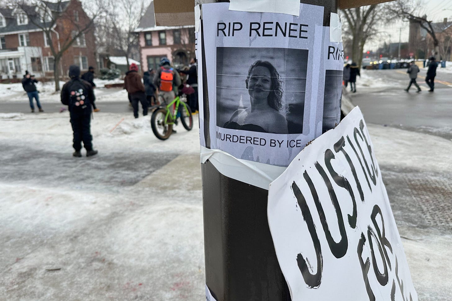 A protest poster taped to a pole reading “RIP RENEE – MURDERED BY ICE,” with people gathered in the background on a winter street.