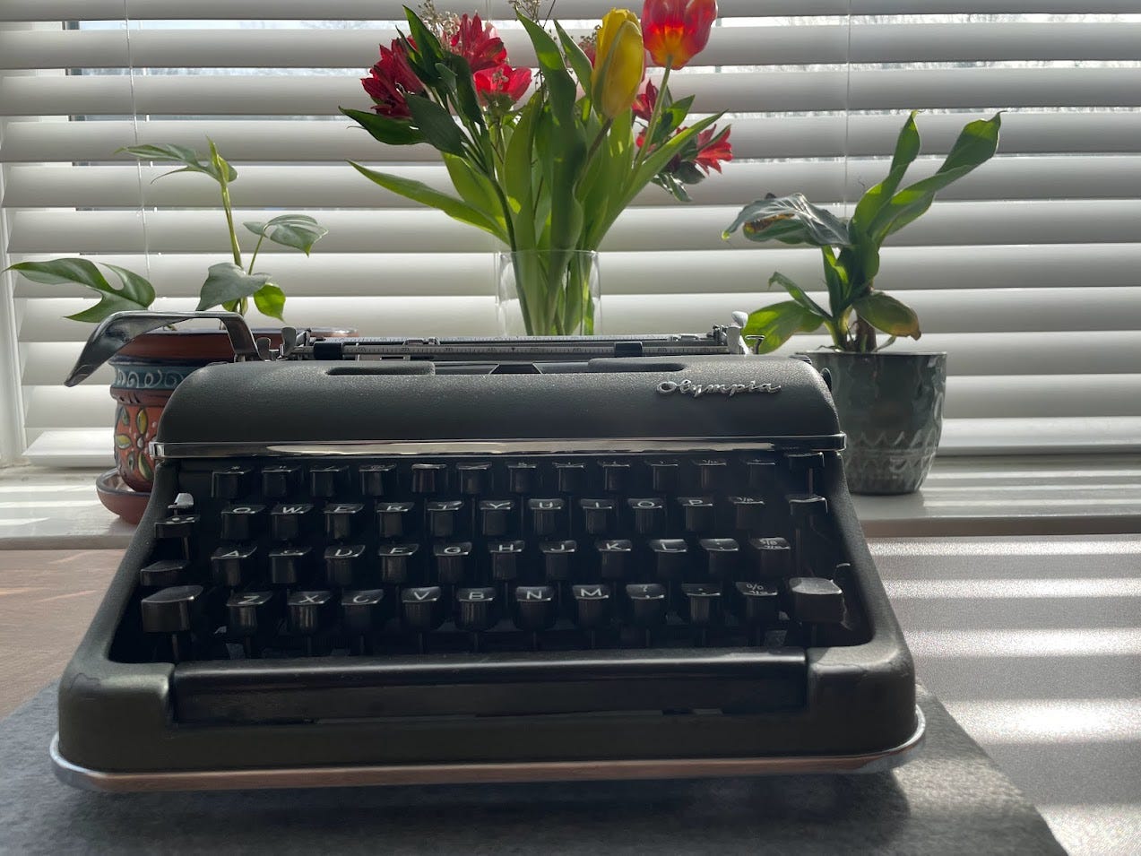 My grey Olympia typewriter on a table in front of a vase of flowers and two houseplants in funky pots, on a sunny day with sunlight streaming through the white blinds.
