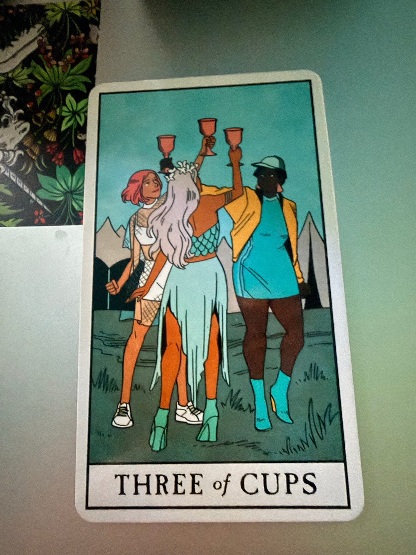 The Three of Cups card from Lisa Sterle’s Modern Witch Tarot, depicting three friends raising goblets together against a turquoise sky and mountain landscape. The Three of Cups card from Lisa Sterle’s Modern Witch Tarot, depicting three friends raising goblets together against a turquoise sky and mountain landscape.