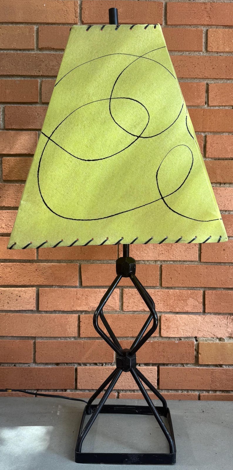 May include: A mid-century modern table lamp with a lime green shade featuring black abstract lines. The shade is stitched along the bottom edge. The lamp has a black metal base with a geometric design and a black cord.