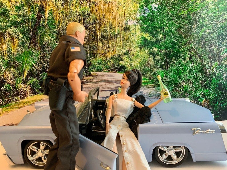 May include: A blue convertible car with a doll in the passenger seat holding a bottle of wine. A police officer stands next to the car.