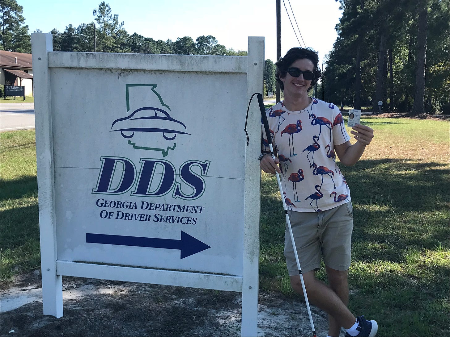 Kai standing next to the Driver Services sign holding his brand new State ID card and his mobility cane. Kai standing next to the Driver Services sign holding his brand new State ID card and his mobility cane.