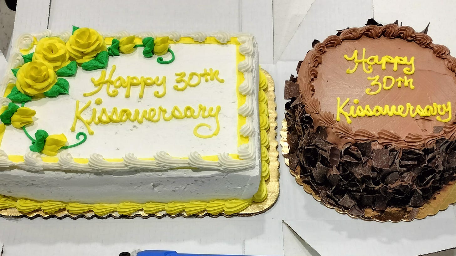 a rectangular white-frosted cake with yellow roses and words, and a chocolate frosted round cake also with yellow frosted words; both cakes read "Happy 30th Kissaversary" a rectangular white-frosted cake with yellow roses and words, and a chocolate frosted round cake also with yellow frosted words; both cakes read "Happy 30th Kissaversary"