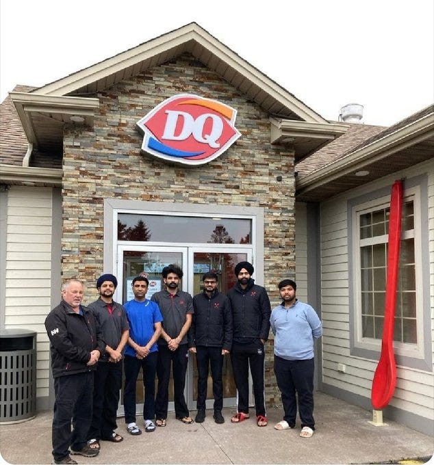 Stone and beige building exterior with large orange and white Dairy Queen logo sign above entrance. Glass door flanked by windows. Red banner hangs on right side. Group of six men stand on concrete pavement in front: leftmost in black jacket and pants with white shoes, next in gray jacket and blue shirt, then blue shirt, black jacket, black jacket and pants with red shoes, light blue shirt and gray pants. All men wear turbans except one with cap. Building has sloped roof with vents.