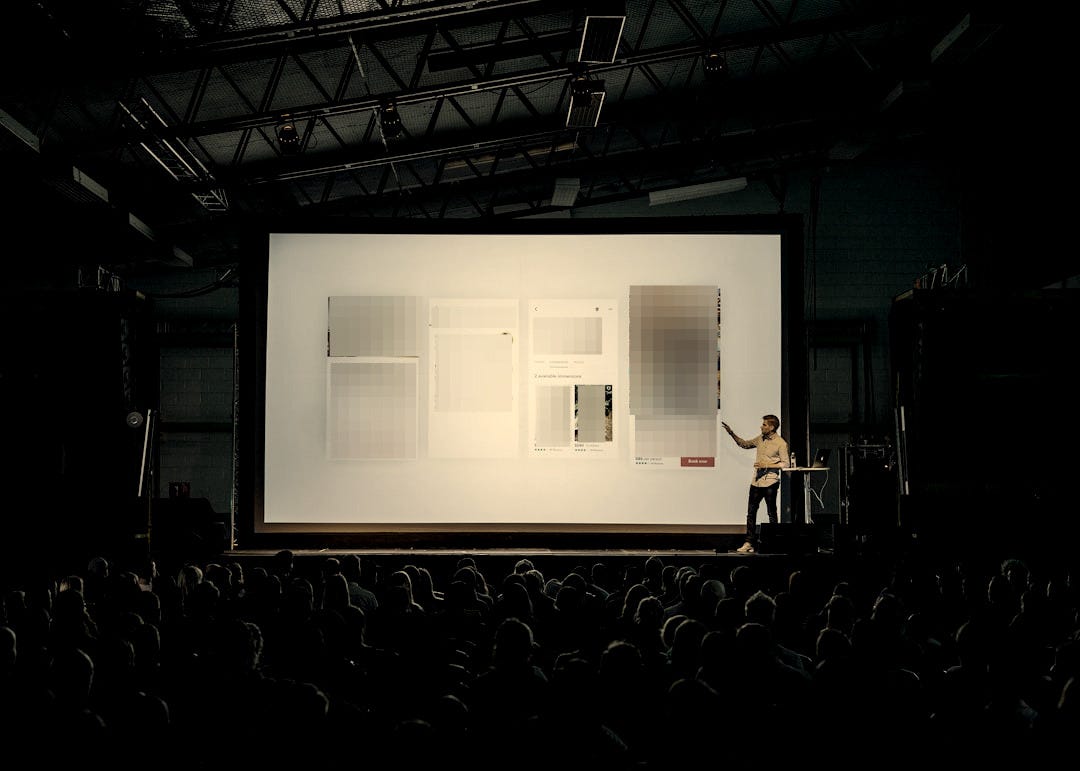 person discussing while standing in front of a large screen in front of people inside dim-lighted room person discussing while standing in front of a large screen in front of people inside dim-lighted room
