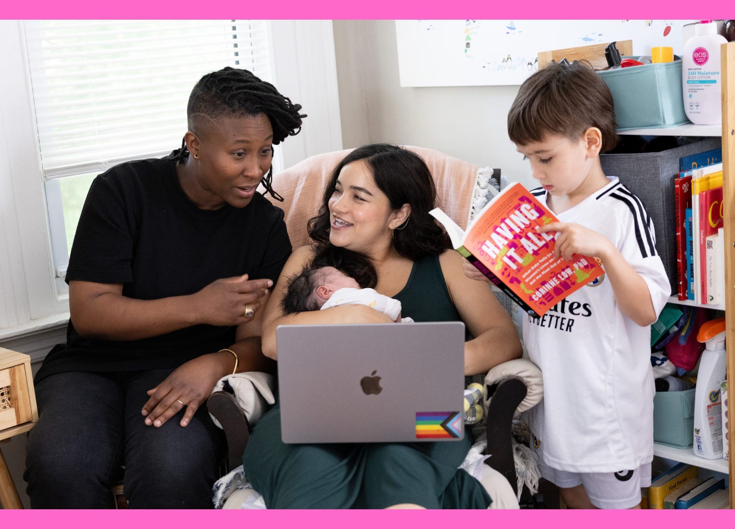 Two women looking at a laptop. One is holding a baby. A little boy reading a book is standing next to the woman with a baby