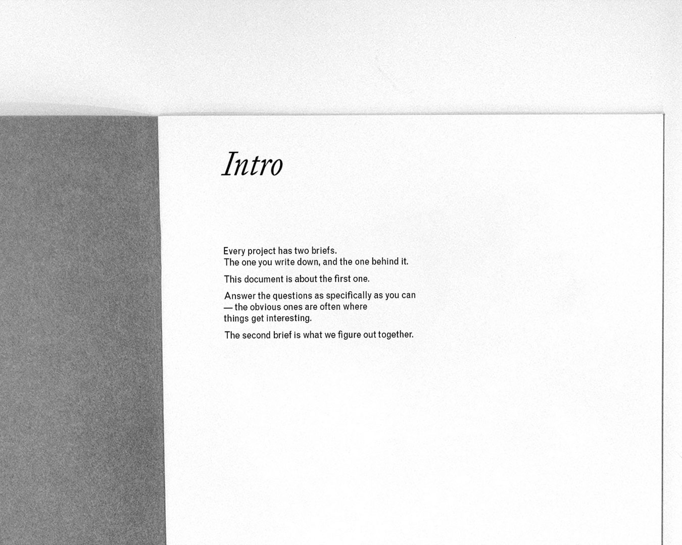 A printed document lies open against a light grey background. The right-hand page is white with the heading Intro in italic serif type. Below, in a smaller sans serif typeface: "Every project has two briefs. The one you write down, and the one behind it. This document is about the first one. Answer the questions as specifically as you can — the obvious ones are often where things get interesting. The second brief is what we figure out together."