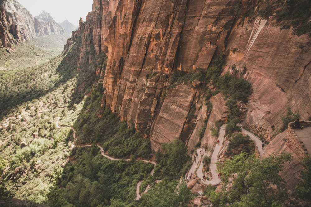A landscape showing a narrow dirt road winding through a tree-filled valley and along the side of red-tinted stone cliffs. A landscape showing a narrow dirt road winding through a tree-filled valley and along the side of red-tinted stone cliffs.
