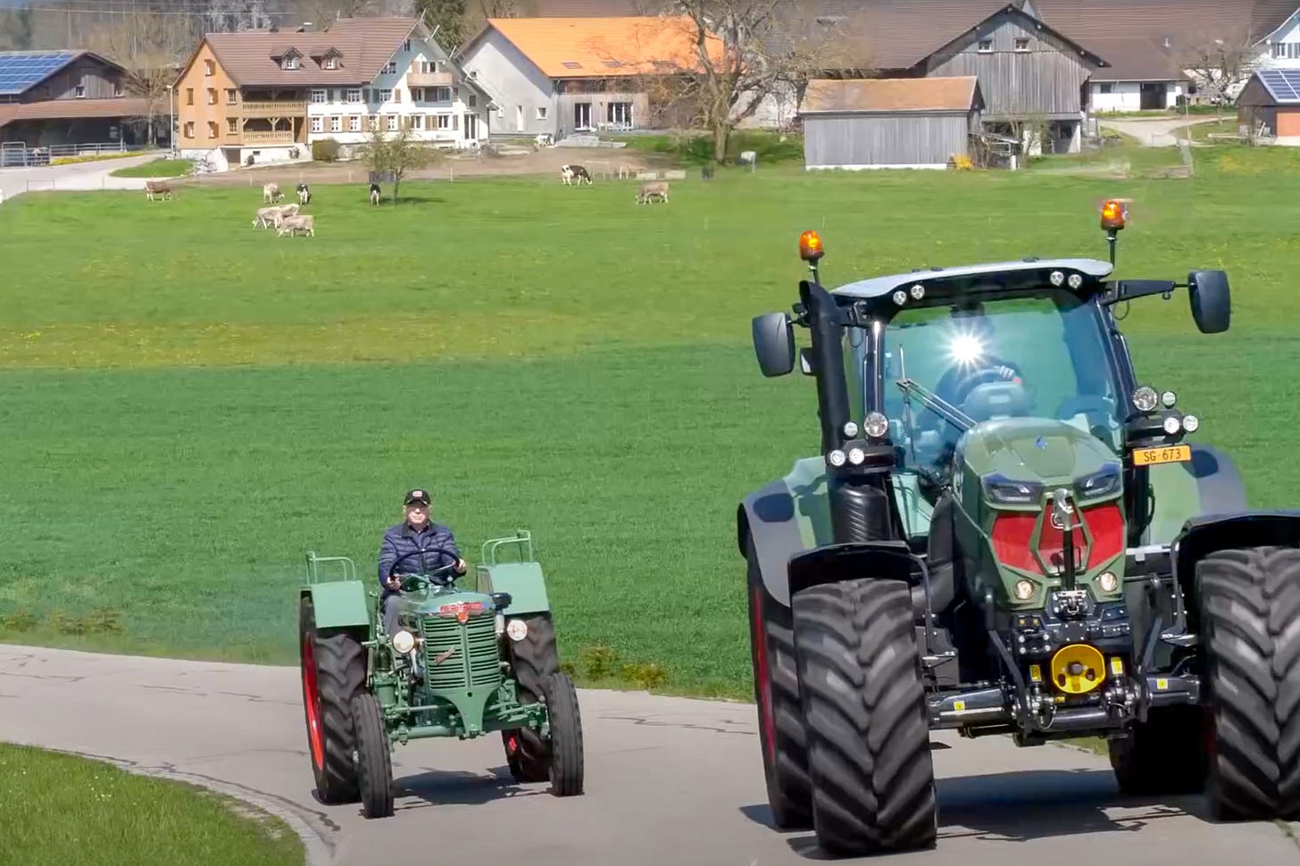 Zwei Hürlimann-Traktoren fahren auf einer schmalen Landstrasse durch grünes Kulturland: Vorne ein grosser moderner Hürlimann-Traktor. Hinten ein kleiner grüner Hürlimann-Oldtimer. An dessen Steuer sitzt Hans Hürlimann Junior, der damals 80jährige Sohn des Firmengründers. Im Hintergrund stehen Bauernhäuser und Ställe, auf der Wiese weiden Kühe. Zwei Hürlimann-Traktoren fahren auf einer schmalen Landstrasse durch grünes Kulturland: Vorne ein grosser moderner Hürlimann-Traktor. Hinten ein kleiner grüner Hürlimann-Oldtimer. An dessen Steuer sitzt Hans Hürlimann Junior, der damals 80jährige Sohn des Firmengründers. Im Hintergrund stehen Bauernhäuser und Ställe, auf der Wiese weiden Kühe.