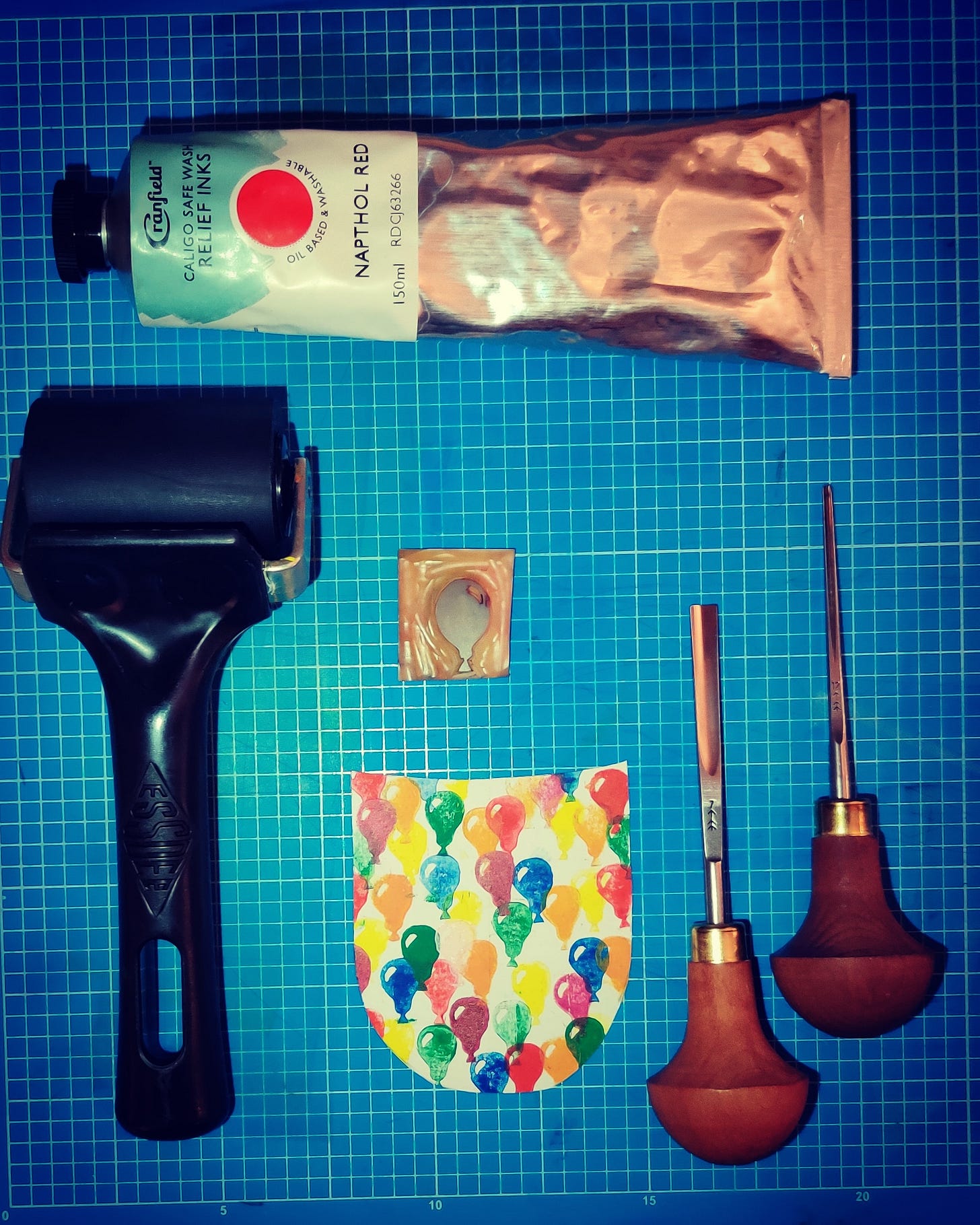 Bright blue cutting mat with white gridlines, with a black print roller, some napthol red ink, two cutting tools, a small piece of carved line and a piece of paper with small balloons printed on it.