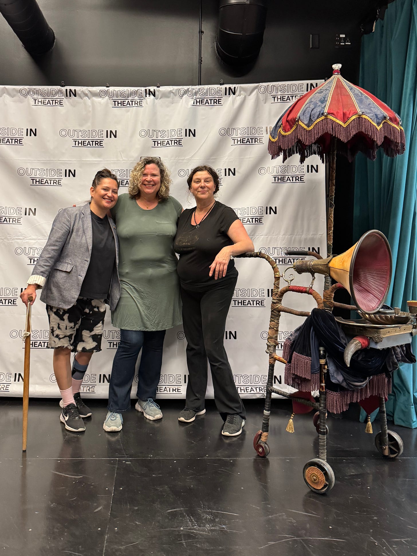 Three smiling women stand in front of a white banner with Outside In Theatre repeated across in. On their right is a walker with attached parasol, silver platter, gramophone horn, and cow horns.
