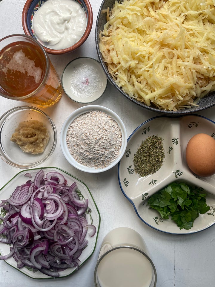 An overhead mise en place for bramboráky: grated potatoes, sliced red onions, whole wheat flour, a cracked egg, fresh parsley, dried marjoram, garlic paste, plant-based milk, sour cream, salt, and a glass of Czech beer.