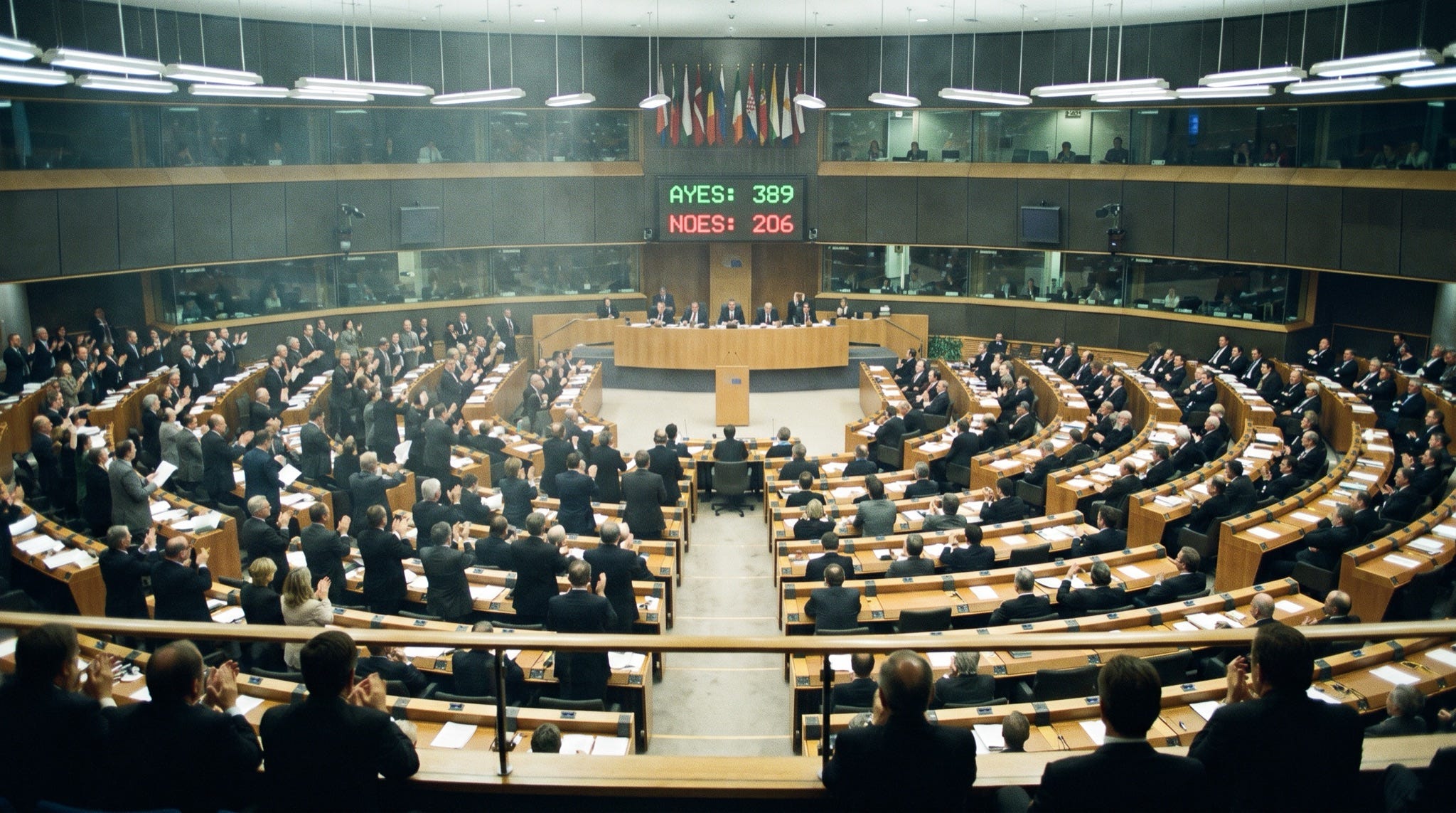A vast parliamentary chamber in the moment after a division, half the delegates standing and applauding while the other half remain seated, the tally board above reading 389 to 206