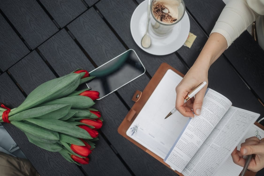 Free A flat lay of an open planner, a bouquet of red tulips, and coffee on a dark table outdoors. Stock Photo