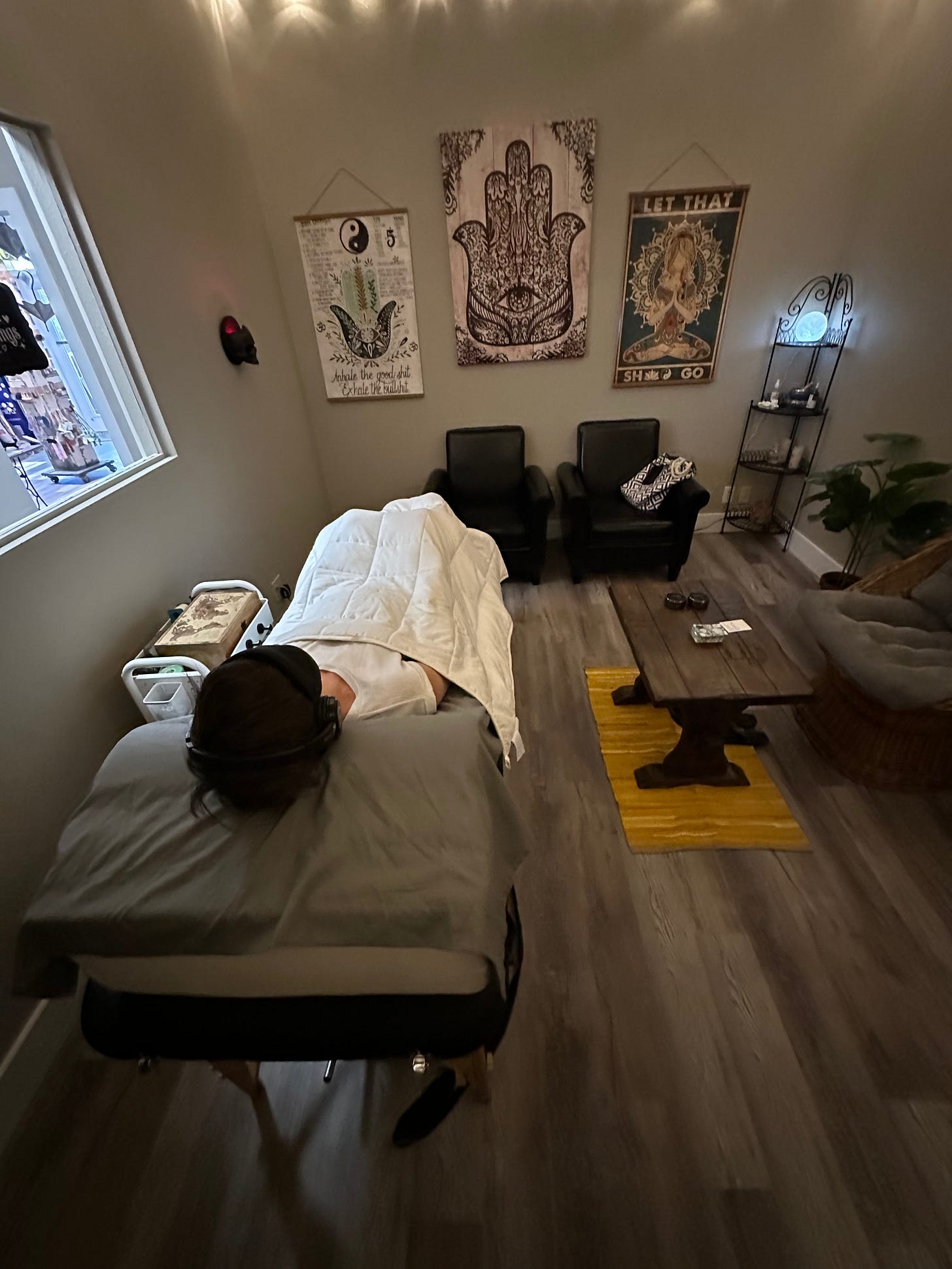 Woman laying on a therapy table, covered in a white blanket. The therapy room has brown walls and boho style furniture and tapestries. Woman laying on a therapy table, covered in a white blanket. The therapy room has brown walls and boho style furniture and tapestries.