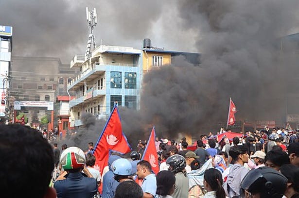 Nepalese citizens protesting in front of a smoking building. Hundreds of people hold flags and face forward to continue marching.