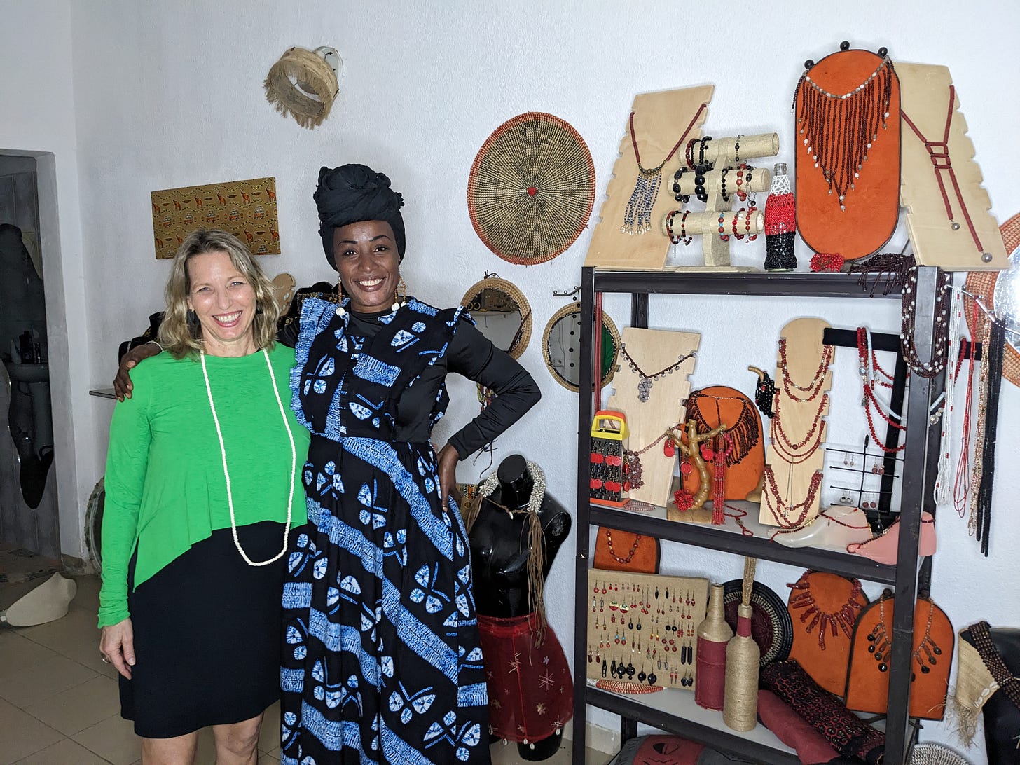 A white woman with blond hair and a black woman with a headwrap are hugging each other and smiling. They are standing in the woman’s jewelry shop. 