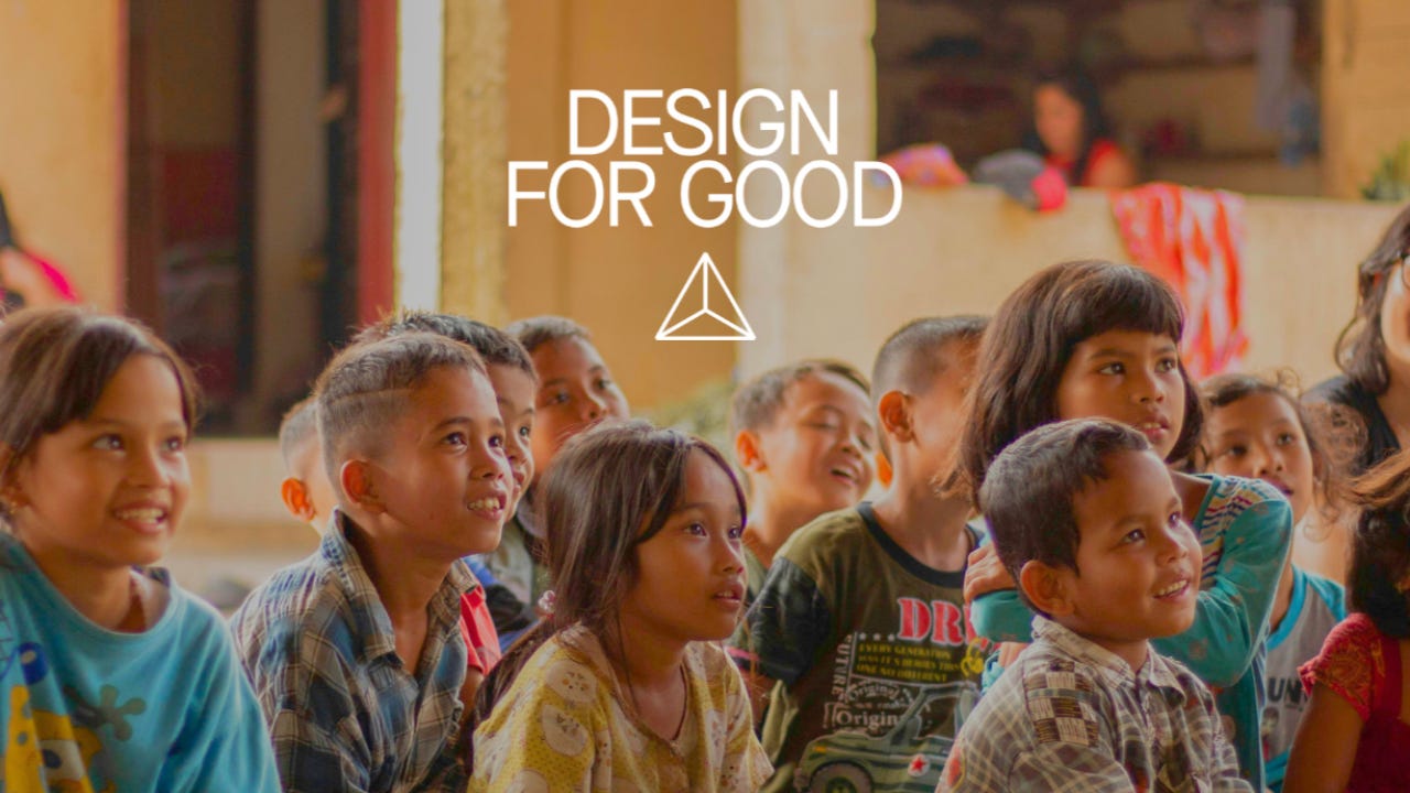 Design for Good logo, white triangle, above about 11 children aged seven sitting on the floor and smiling, looking at someone off-picture, probably a teacher or guest telling them something cool.