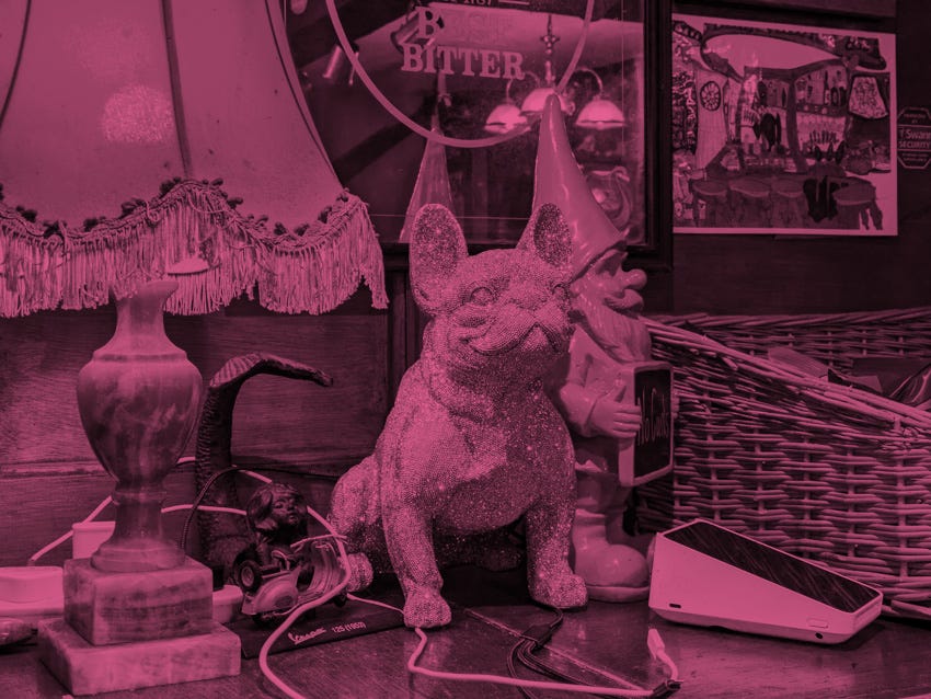 Clutter on the back bar of The Swan With Two Necks, including a gnome, a glitter-covered toy dog, a card machine, and an old lamp.