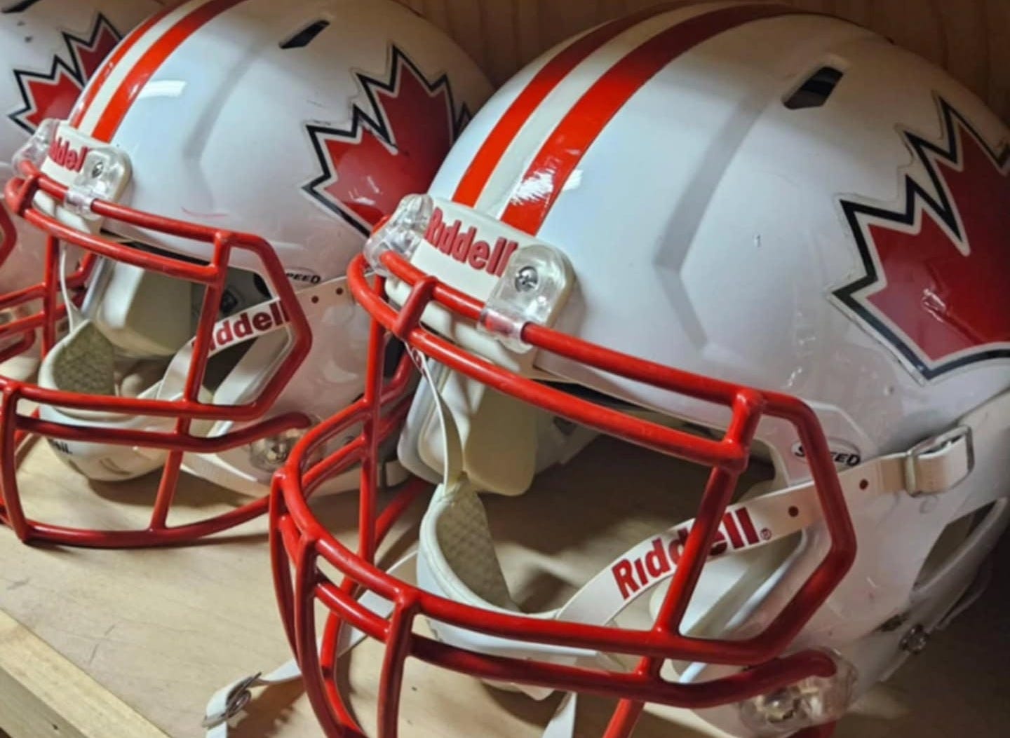 Three white and red helmets, with red Canadian maple leafs on the side of the head and red facemasks, sit on a wooden shelf. Three white and red helmets, with red Canadian maple leafs on the side of the head and red facemasks, sit on a wooden shelf.