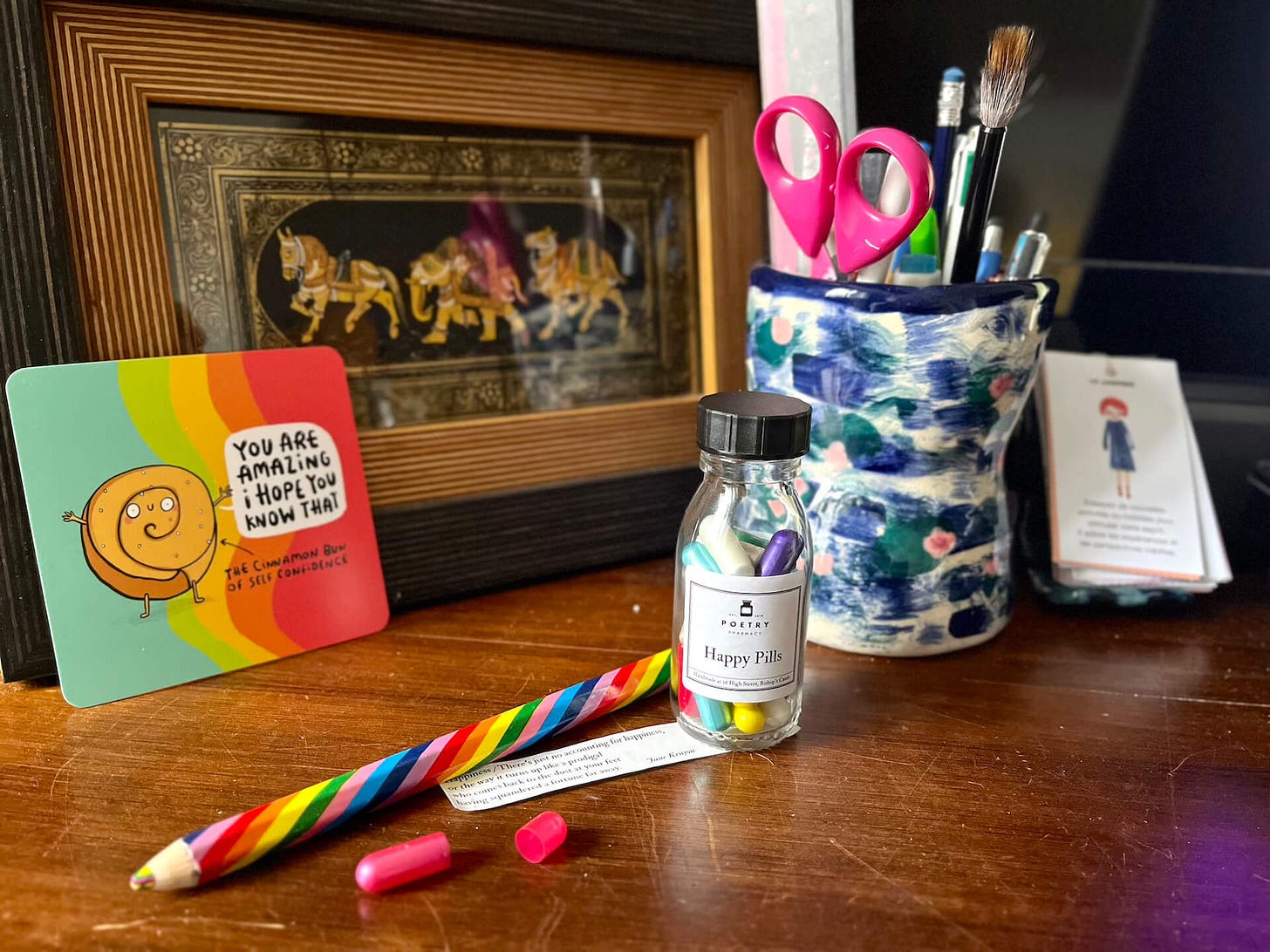 Photo of Claire's desk featuring indian art of a horse elephant and camel framed in the back, a ceramic pot she created on the right, an oracle card on the left that says you are amazing i hope you know that from the cinnamon bun of self confidence, and a happy pills glass bottle from the poetry pharmacy center with a rainbow pencil next to it and a quote on happiness in the paper in one of the pill cases
