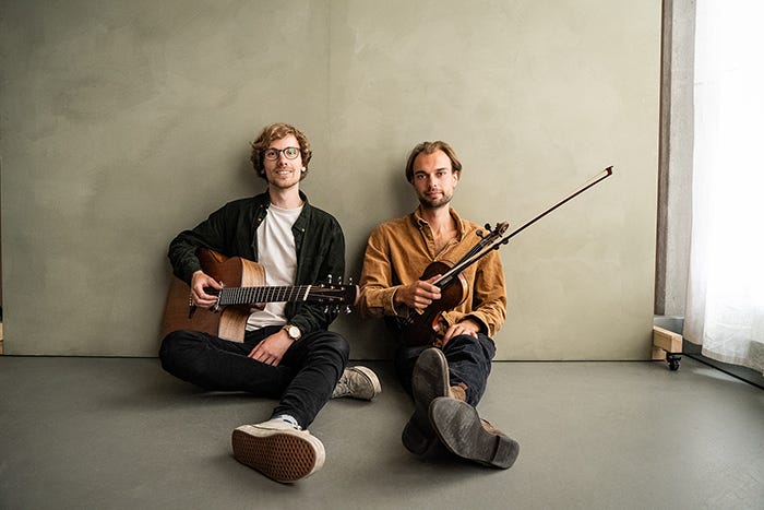 Filkin's Drift - two men, one with a fiddle, the other holding a guitar, sit on a bare floor, their backs against a wall. Filkin's Drift - two men, one with a fiddle, the other holding a guitar, sit on a bare floor, their backs against a wall.