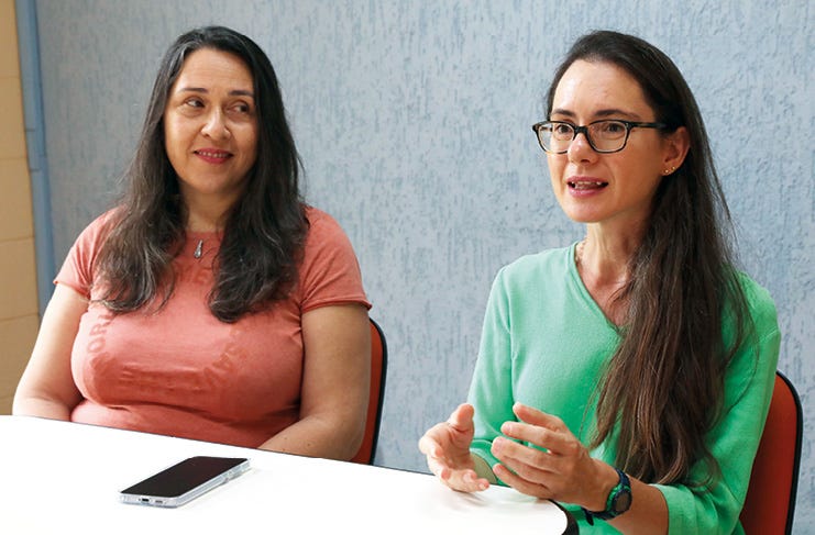 Duas pessoas de cabelos longos e escuros sentadas lado a lado em uma mesa branca, sendo que uma veste camiseta rosa e a outra usa óculos de armação escura e blusa verde, aparentando estar em uma conversa ou reunião, com um smartphone apoiado sobre a mesa. Duas pessoas de cabelos longos e escuros sentadas lado a lado em uma mesa branca, sendo que uma veste camiseta rosa e a outra usa óculos de armação escura e blusa verde, aparentando estar em uma conversa ou reunião, com um smartphone apoiado sobre a mesa.