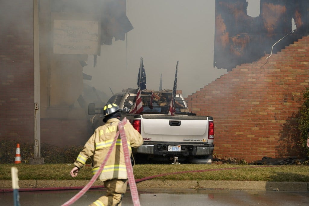 A firefighter walks towards the Church of Jesus Christ of Latter-day Saints which is on fire, with a white pickup truck parked nearby.