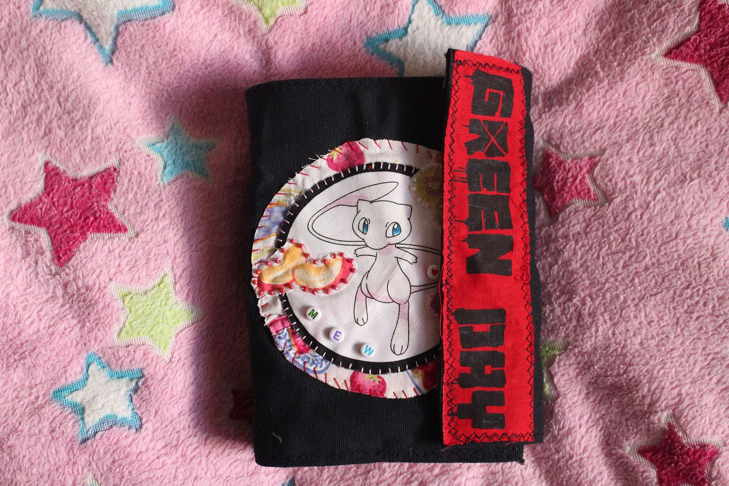 a black, clothbound sketchbook with a patch of the pokemon "Mew" sewn to the front, with a flap covering it. the flap has a red and black Green Day patch sewed to it