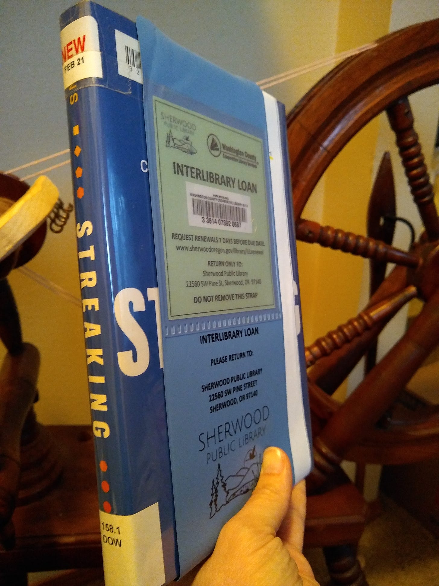 A hand holding the book Streaking, with an Interlibrary Loan band on it, in front of a spinning wheel