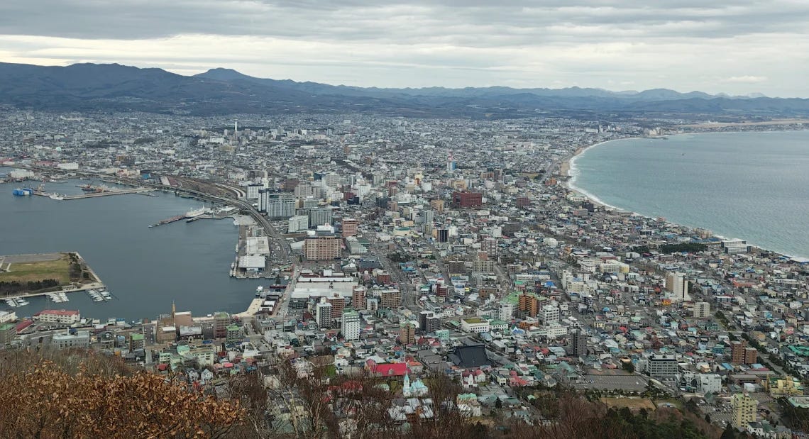 A panoramic view of the city of Hakodate from the summit of Mt. Hakodate on a cloudy day, showing the city's unique shape, the busy harbor on the left, and the open ocean on the right. A panoramic view of the city of Hakodate from the summit of Mt. Hakodate on a cloudy day, showing the city's unique shape, the busy harbor on the left, and the open ocean on the right.