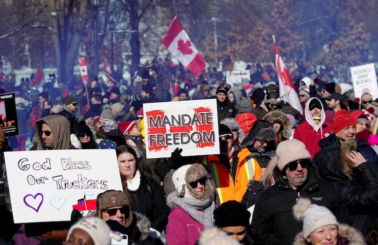 A crowd of people holding signs. One visible sign says 'MANDATE FREEDOM' and another says 'God Bless Our Truckers.' A crowd of people holding signs. One visible sign says 'MANDATE FREEDOM' and another says 'God Bless Our Truckers.'