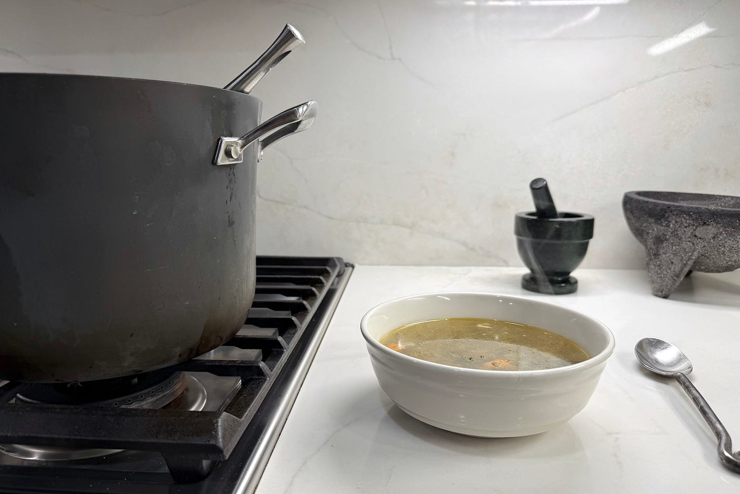 Stockpot on the stove beside a warm bowl of soup with a spoon on the counter.” Stockpot on the stove beside a warm bowl of soup with a spoon on the counter.”
