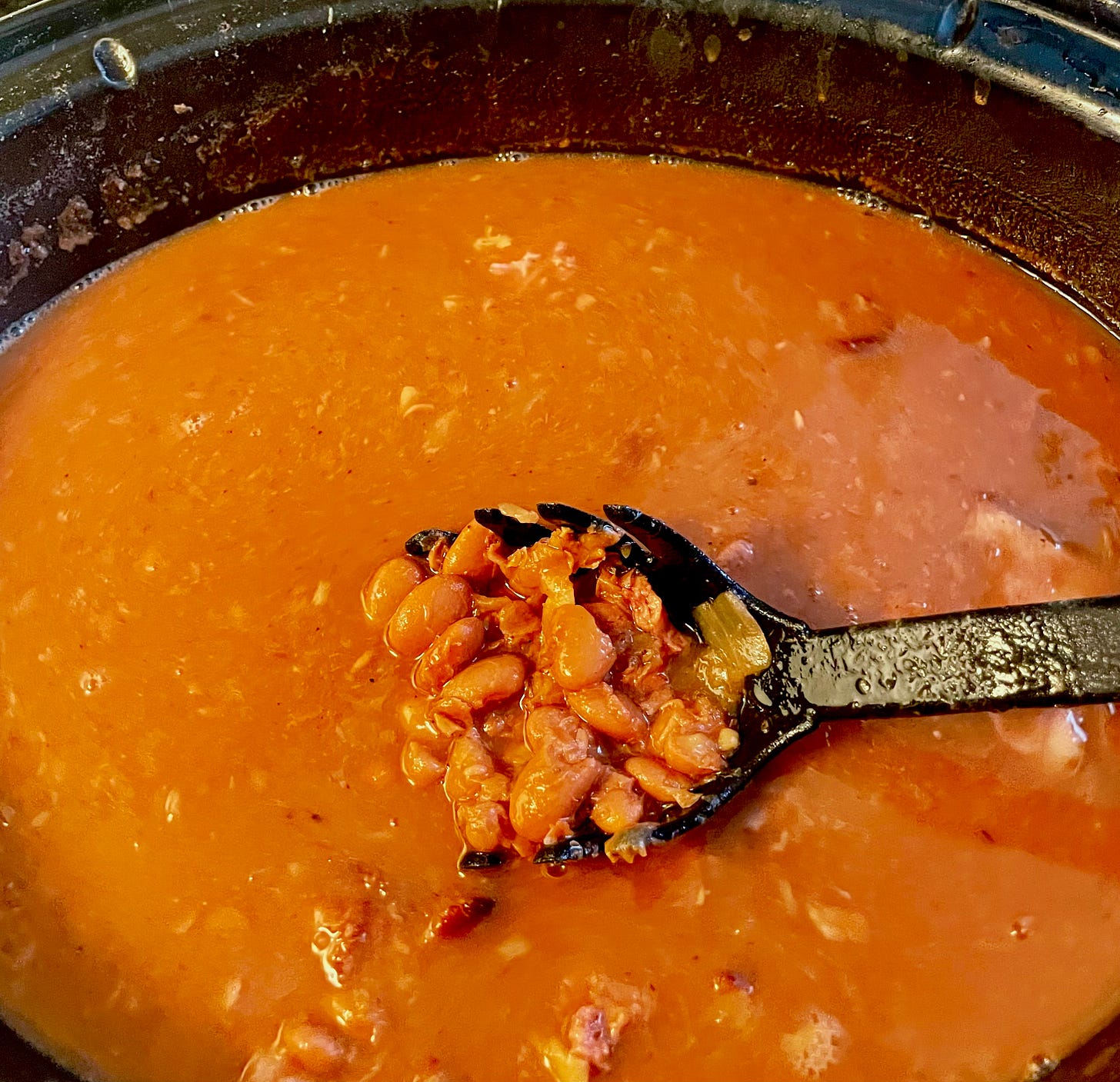 In a black crockpot, close up of the bean juice and one slotted spoonful of beans suspended over it