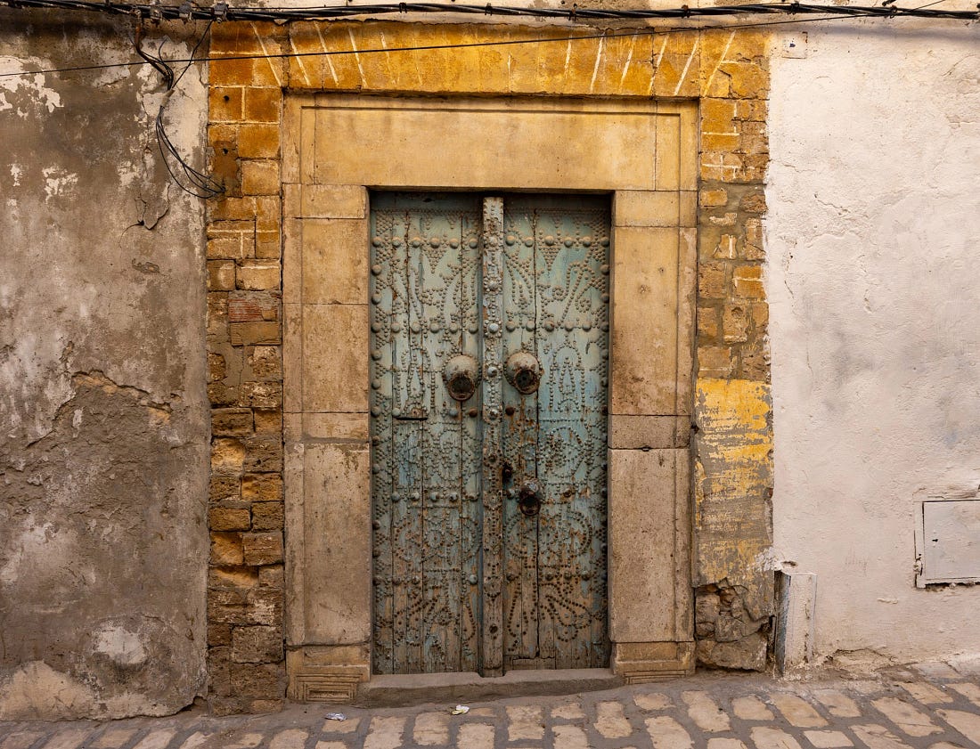 Weathered studded wooden door set into a stone frame on Rue Meffrej in the Tunis Medina, showing layers of age, repair, and daily use.