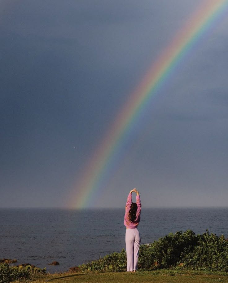 This may contain: a woman standing on top of a lush green field next to the ocean under a rainbow