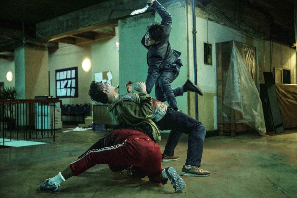 three people fighting; one bent backwards over a guy in a red tracksuit, one jumping up to stab him with a knife. background is dilapidated interio