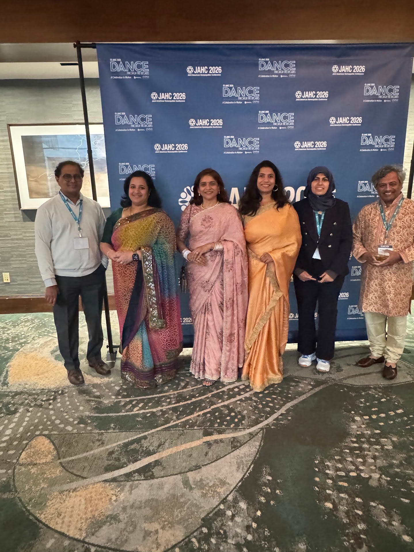 Six conference attendees, several in colorful saris, pose at the JAHC 2026 Dance Through the Decades awards dinner backdrop. 