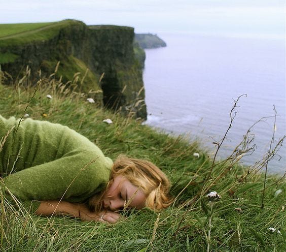 This may contain: a woman laying on top of a lush green hillside next to the ocean with her eyes closed