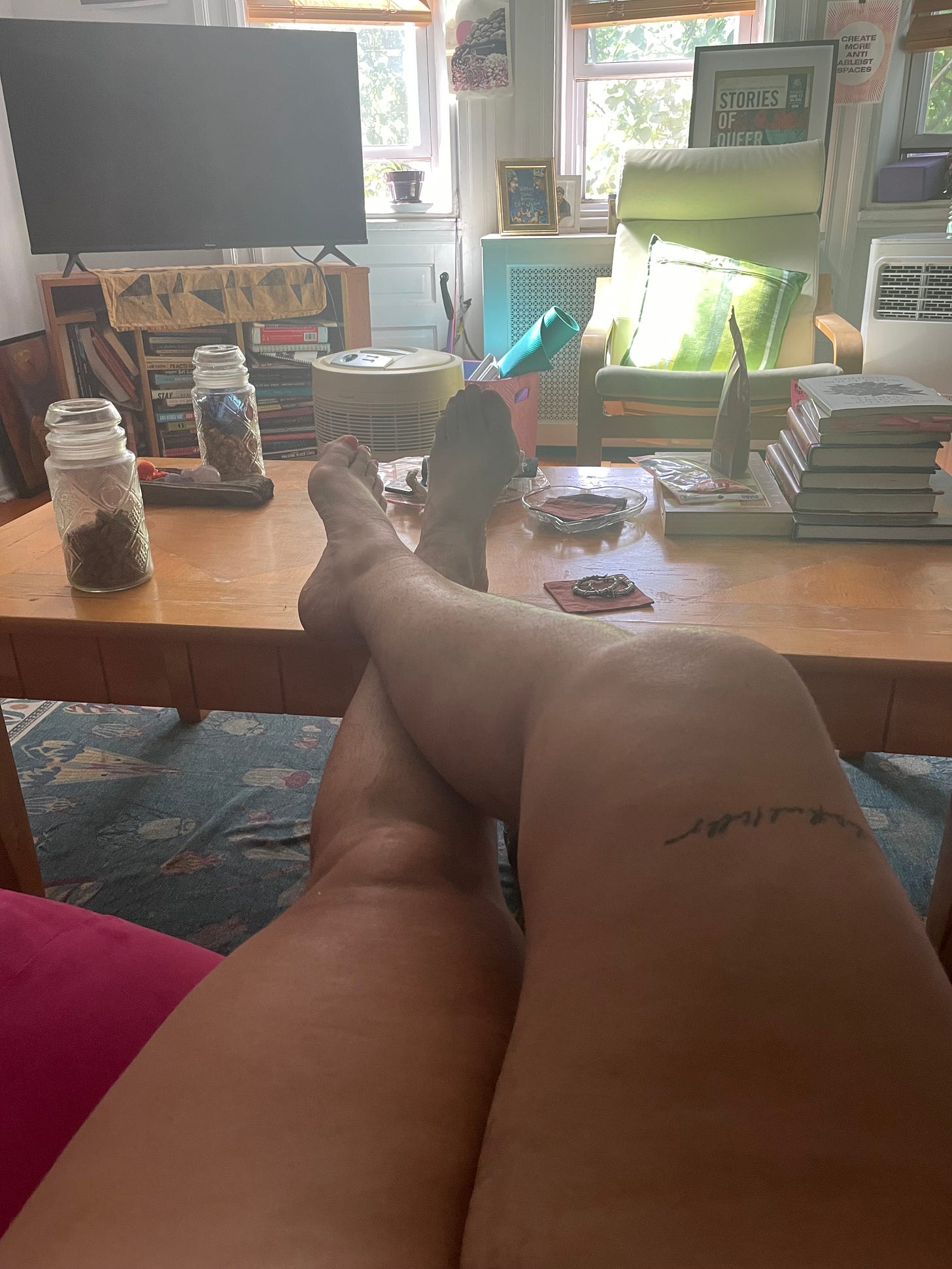 the author's thick thighs and calves crossed, resting on a wooden coffee table stacked with books, jars of candy, driftwood, a crystal ashtray with costers, a tv and posters are in the background, you can feel the haze of the heat of a 103 degree heat dome day the author's thick thighs and calves crossed, resting on a wooden coffee table stacked with books, jars of candy, driftwood, a crystal ashtray with costers, a tv and posters are in the background, you can feel the haze of the heat of a 103 degree heat dome day