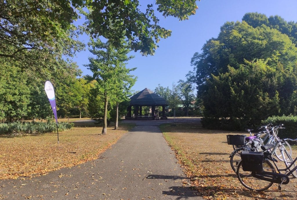 Two bikes and the parkrun flag stand next to a tarmacced path leading to the pavilion, which is an open air structure with a leaning roof in the middle of a tarmacced area.