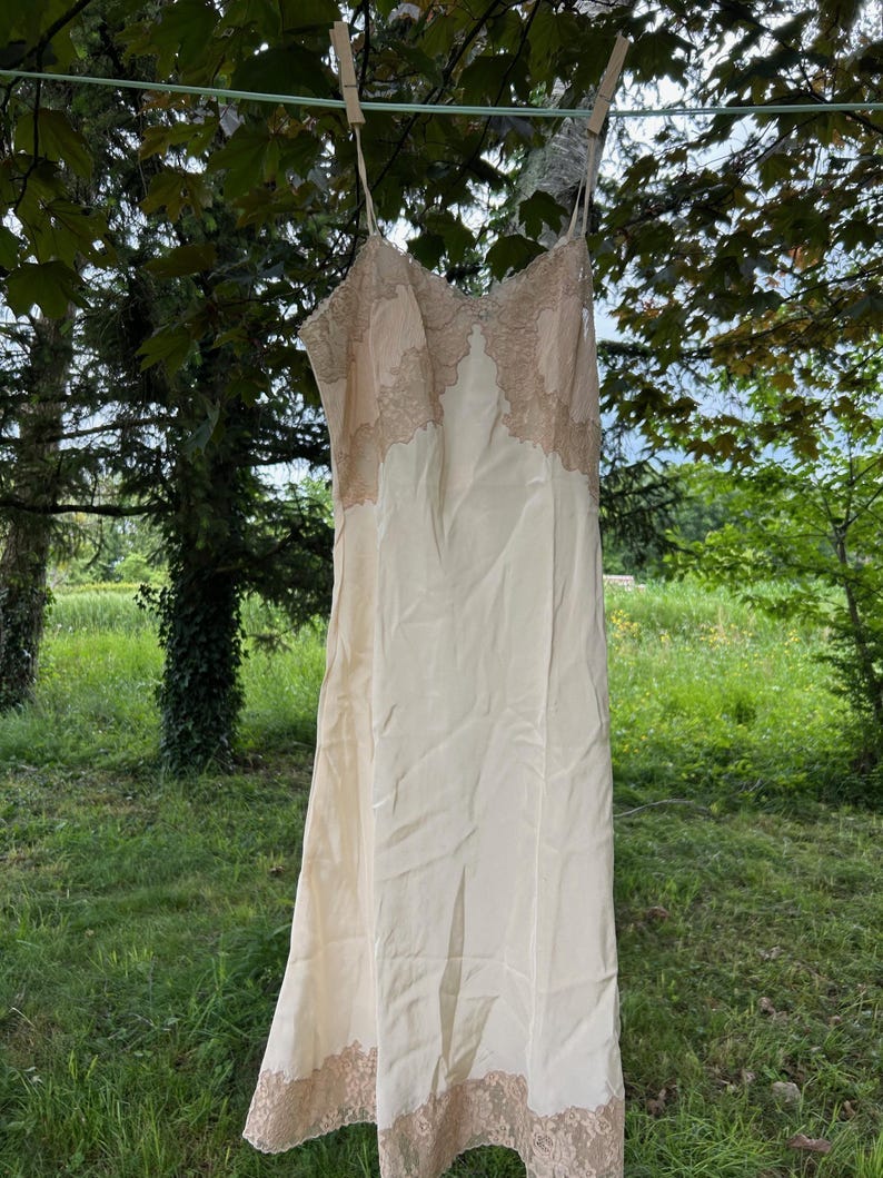 May include: A cream-colored slip dress with delicate lace trim along the neckline and hemline, hanging on a clothesline. The dress has thin straps and a flowing silhouette, with a subtle sheen. The lace is a light tan color.