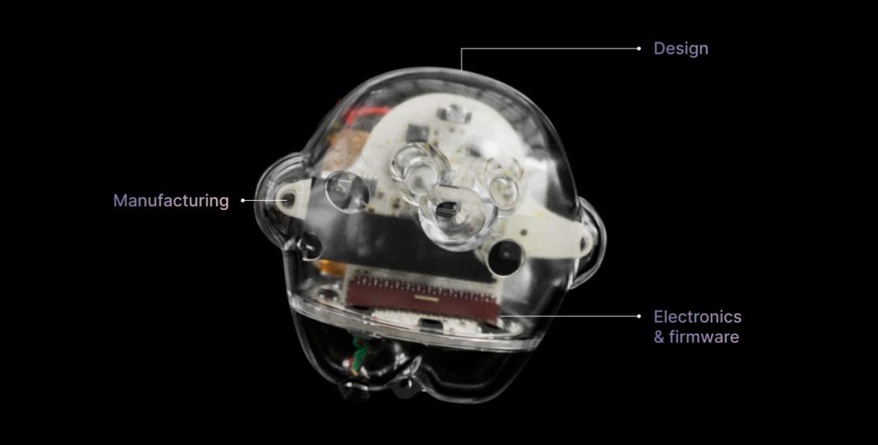 Transparent robotic head or skull showing internal components with three labeled elements: "Design" pointing to the outer form, "Manufacturing" indicating the mechanical assembly and gears, and "Electronics & firmware" highlighting the circuit board and electronic components inside.